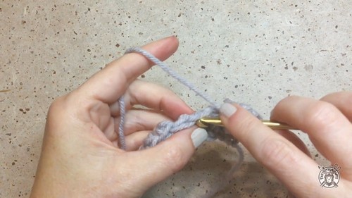 Starting the foundation chain for the grey crochet sweater vest body with a gold hook on a stone surface.