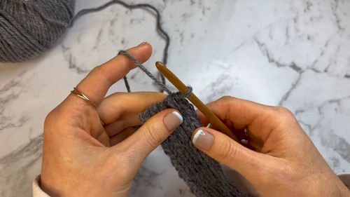 Hands building a grey crochet ribbing foundation on a gold hook against a marble surface with a yarn ball in the background