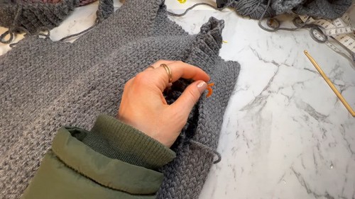 Hand placing a stitch marker at the armhole edge of a flat grey crochet vest panel laid on a marble surface