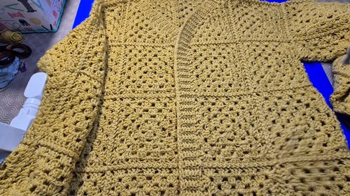 YouTube screenshot showing a completed mustard yellow granny stitch sweater laid flat on a blue surface with visible panel seams and ribbed cuffs
