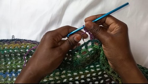 Hands working a filet mesh row in multicolor variegated yarn with a blue hook along the top edge of a crochet panel.