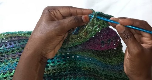 Hands joining a seam along the curved edge of a multicolor variegated filet crochet panel with a blue hook.