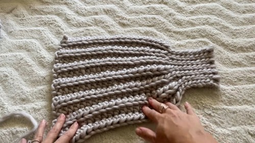 Hands smoothing a shaped chunky grey ribbed crochet sweater body panel flat on a white textured surface showing the tapered silhouette