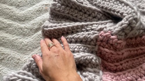 Hand resting on a chunky crochet ribbed fabric panel in grey with pink edge visible, showing the deep vertical rib texture of the post stitch pattern