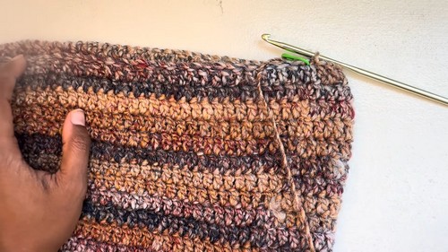 Hand holding a completed crochet sweater body section in multicolor variegated yarn with a green hook resting at the top edge against a white wall