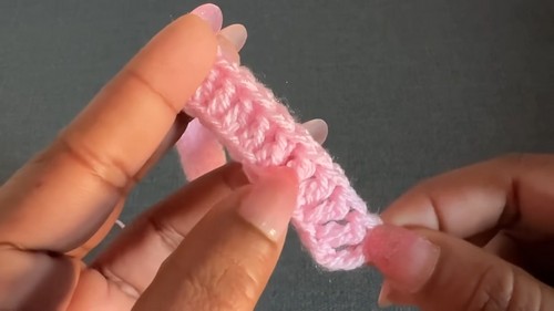 Fingers holding a short pink foundation chain of the first few stitches in pale pink yarn, plain grey background