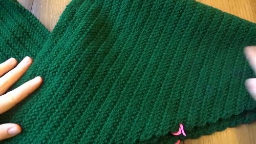 Two flat green crochet panels laid side by side on a wooden surface showing the diagonal wrap front shaping with a pink stitch marker