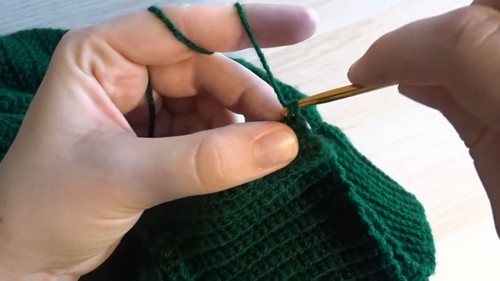 Hands using a gold crochet hook to work stitches into the edge of a forest green ribbed crochet wrap sweater panel