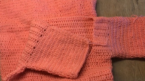 Coral crochet garment panels laid flat showing the mesh body and a ribbed sleeve cuff in progress