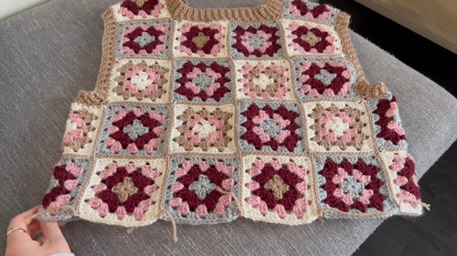 Completed granny square crochet vest panel in burgundy, pink, gray, and tan laid flat on a gray sofa before assembly.