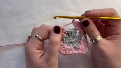 Hands working the second round of a small pink and gray granny square with a gold hook on a white surface.