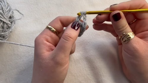Hands crocheting the center magic ring of a gray and pink granny square with a gold hook on a white surface.