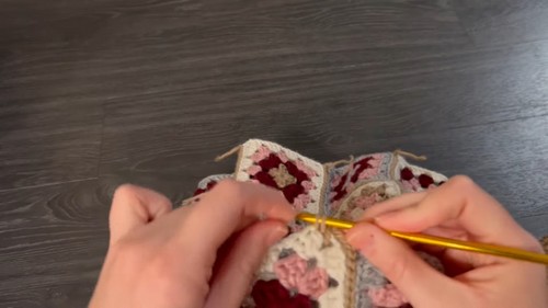Hands joining two rows of multicolor granny squares with a gold hook on a dark wood surface.