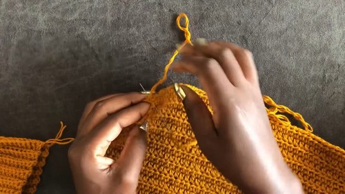 Hands guiding a gold hook through the shoulder increase point of a mustard yellow crochet raglan sweater in progress.