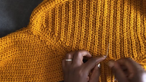 Hands using a tapestry needle to seam the underarm join of a mustard yellow ribbed crochet raglan sweater.