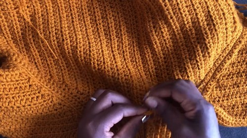 Hands weaving in a yarn tail at the center of a completed mustard yellow crochet turtleneck sweater laid flat.