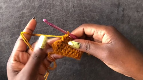 Hands working the first rows of a mustard yellow ribbed crochet band with a pink hook against a gray background.