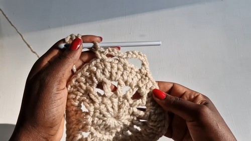 Hands working the outer round of a large circular cream crochet motif with a white hook.