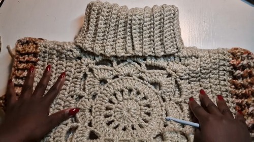 Hands smoothing a large cream granny square panel flat with a folded ribbed turtleneck collar visible above it.