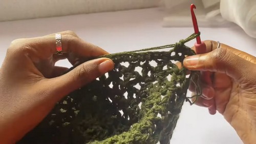 Hands crocheting an olive green lace sweater panel with a red hook, open star stitch texture visible across the fabric