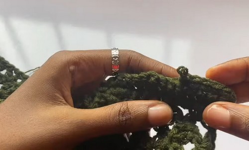 Close view of hands holding a partially worked olive green crochet fabric showing dense cluster texture along a finished edge