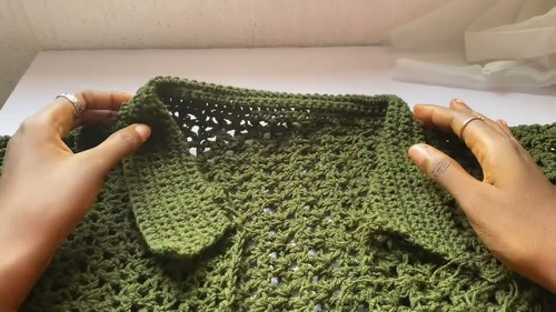 Hands holding a partially completed olive green crochet sweater open to show the ribbed collar, front opening and lace body panels laid flat