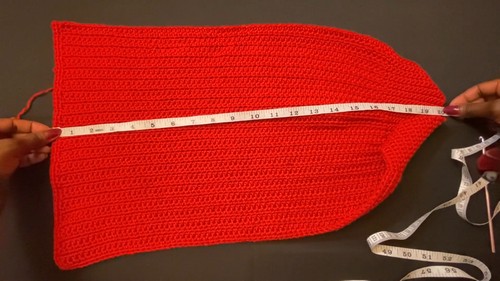 Hands holding a measuring tape across the width of a red ribbed crochet sweater panel on a dark surface.