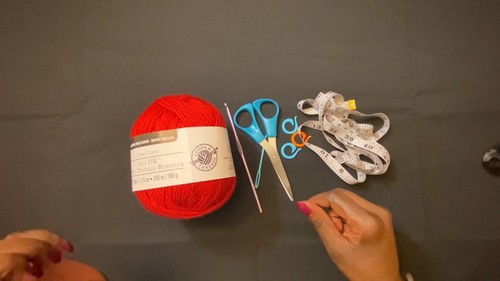 Red yarn ball with label, blue and silver scissors, a small crochet hook, and a measuring tape laid out on a dark surface.