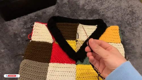 Hands holding the black V-neckline edging being added to a partially assembled patchwork crochet sweater vest laid flat on a carpet