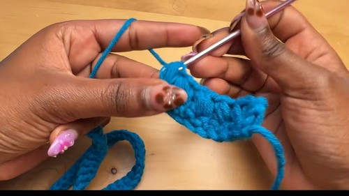hands forming the beginning of a puff stitch in teal blue yarn using a silver hook on a wooden surface