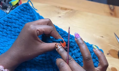 hands working a purple crochet hook through teal blue puff stitch fabric, orange stitch marker clipped to the work