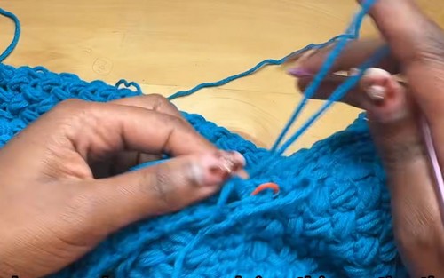 close-up of hands pulling multiple yarn loops through teal blue crochet fabric, orange stitch marker visible mid-work