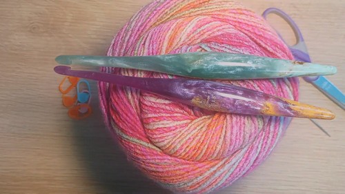 Ball of pink and orange variegated yarn with two crochet hooks and stitch markers on a wooden surface