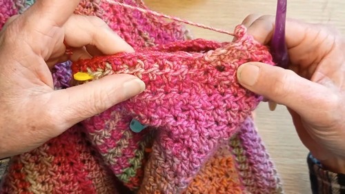 Hands with stitch markers holding the raglan shaping section of a pink and orange variegated crochet sweater in progress