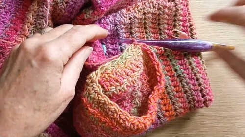 Hands using a purple hook to pick up stitches along the armhole edge of a multicolor crochet sweater body