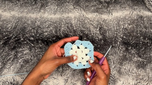 Hands holding one finished granny square in light blue, cream and black on a grey fur blanket