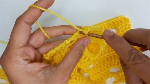 Hands guiding yellow yarn through the open corner of a lace panel with a wood hook.