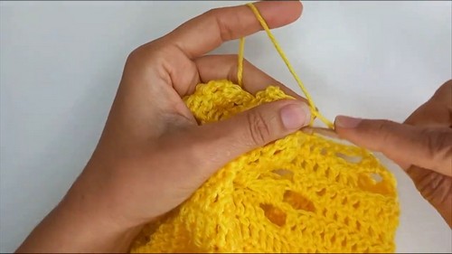 Hands shaping a yellow lace motif cluster with fingers while pulling yarn through with a wood hook.