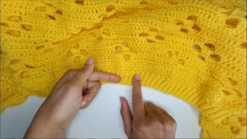 Hands pointing to the transition between the floral eyelet and solid crochet sections of a yellow sweater laid flat.