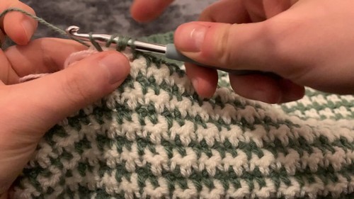 close-up of hands working a silver hook through alternating green and cream houndstooth stitch rows