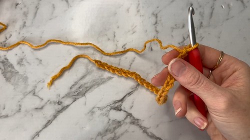 Hand holding a red crochet hook with a short foundation chain in mustard yarn on a marble surface