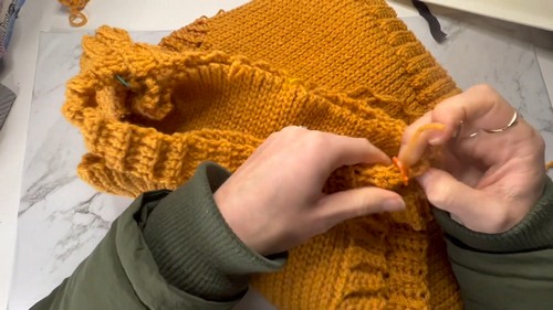 Hands attaching the ribbed armhole edging strip to the body of a mustard yellow crochet vest on a marble surface