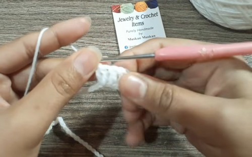 Hands working the first white stitches of a crochet collar with a pink hook on a wooden table