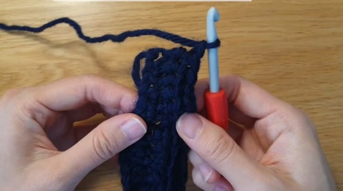 Hands holding the beginning rows of a navy ribbed crochet swatch with a red-handled hook, foundation chain visible at the top