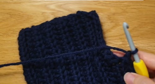 Navy ribbed crochet sweater body panel laid flat on a wood surface with a grey hook resting at the upper right corner