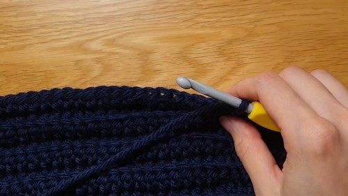Hand using a yellow-handled grey crochet hook along the top edge of a completed navy ribbed crochet sweater body panel on a wood surface