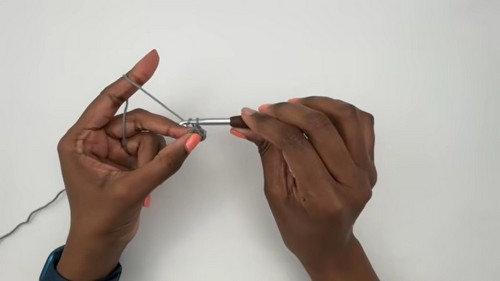 Hands forming the beginning chain loop with grey yarn and a silver crochet hook against a white background