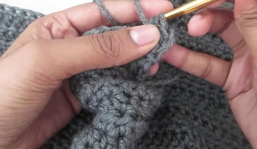 Grey crochet body fabric being worked in rows with a gold hook showing grouped stitches