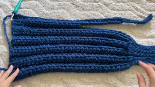 hands stretching a wide navy blue ribbed crochet panel flat on a white surface, green hook visible at left edge