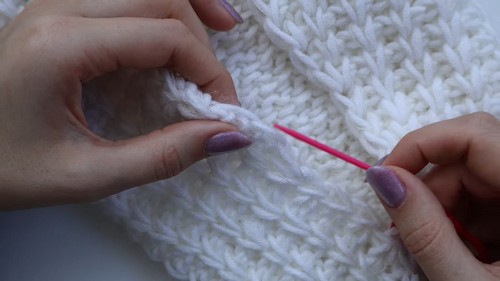 Hands using a pink crochet hook to work stitches into a dense white Tunisian crochet fabric showing a waffle-like grid texture